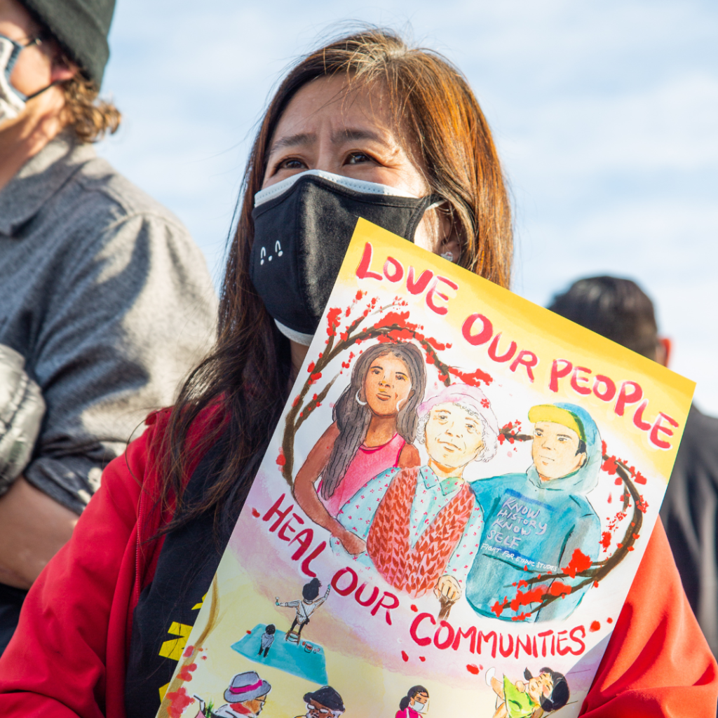 Image of a masked woman at a protest. She is holding a sign that says, "Love our people, heal our communities."